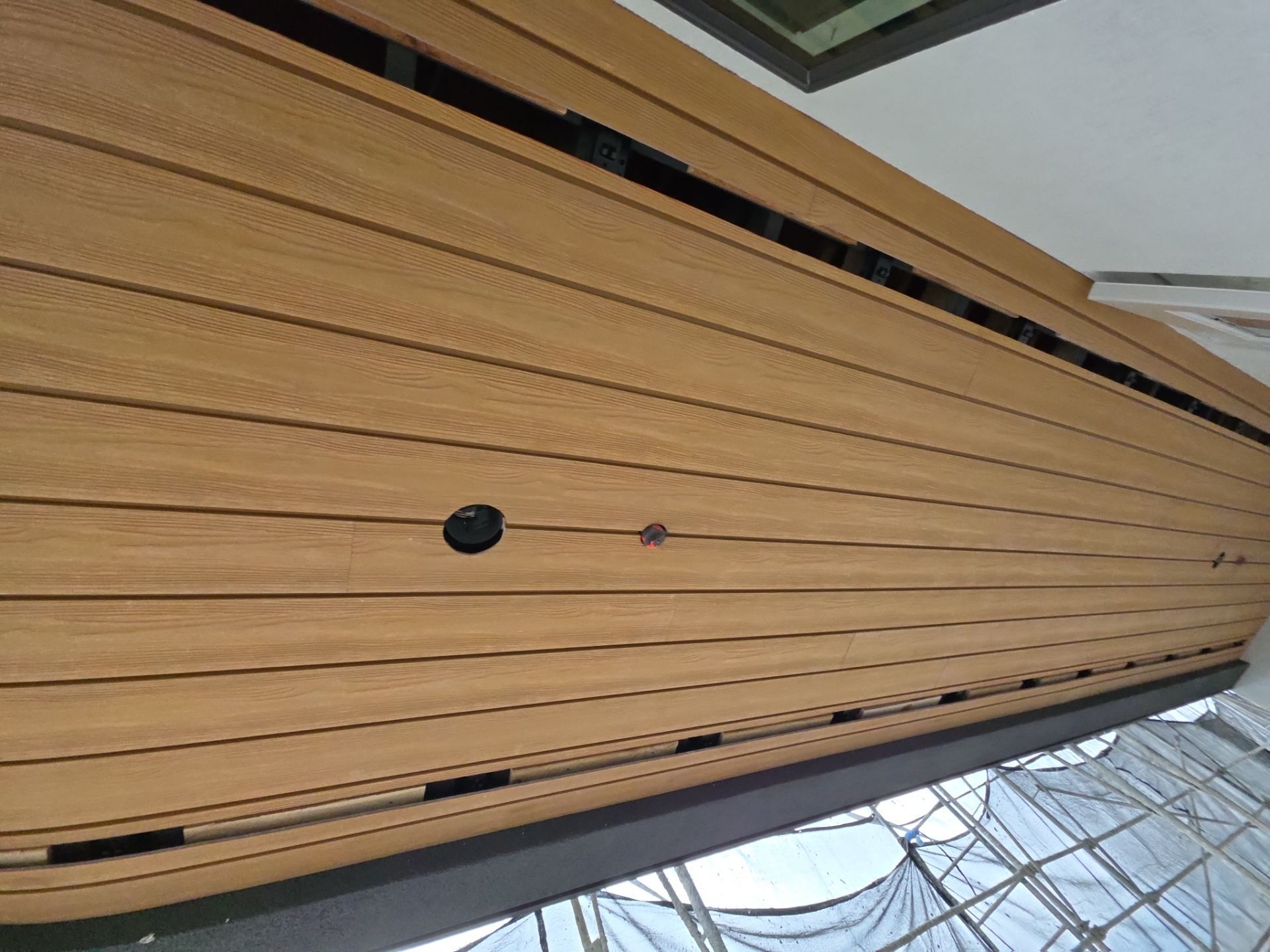 Wood-paneled wall with a round black fixture, viewed at an angle indoors near a window.