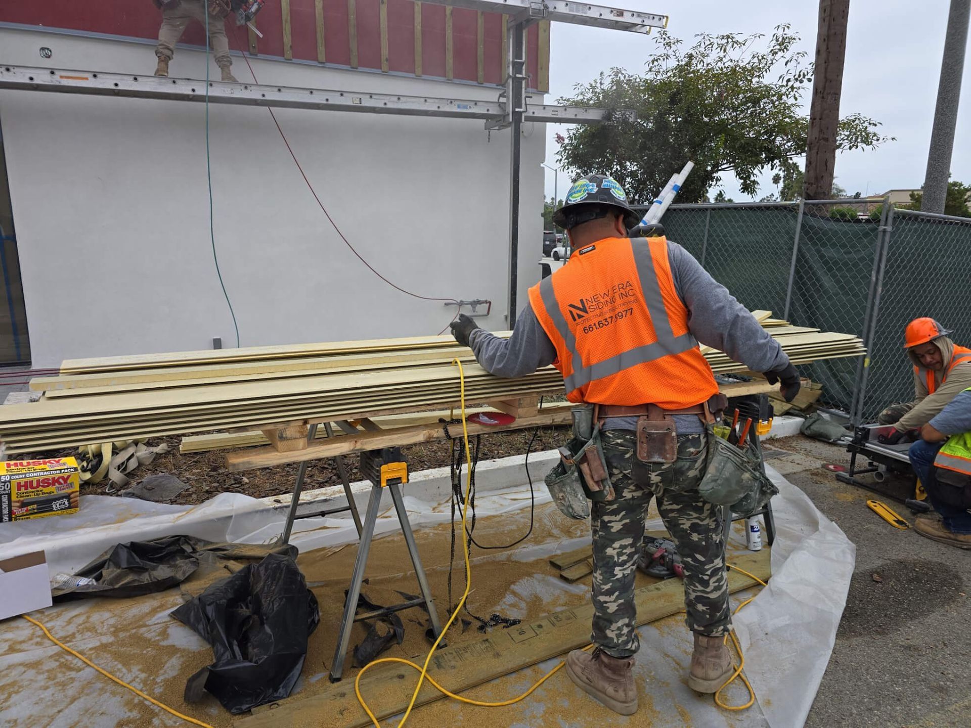 Construction workers in safety vests work outdoors at a job site, painting wood boards on sawhorses.
