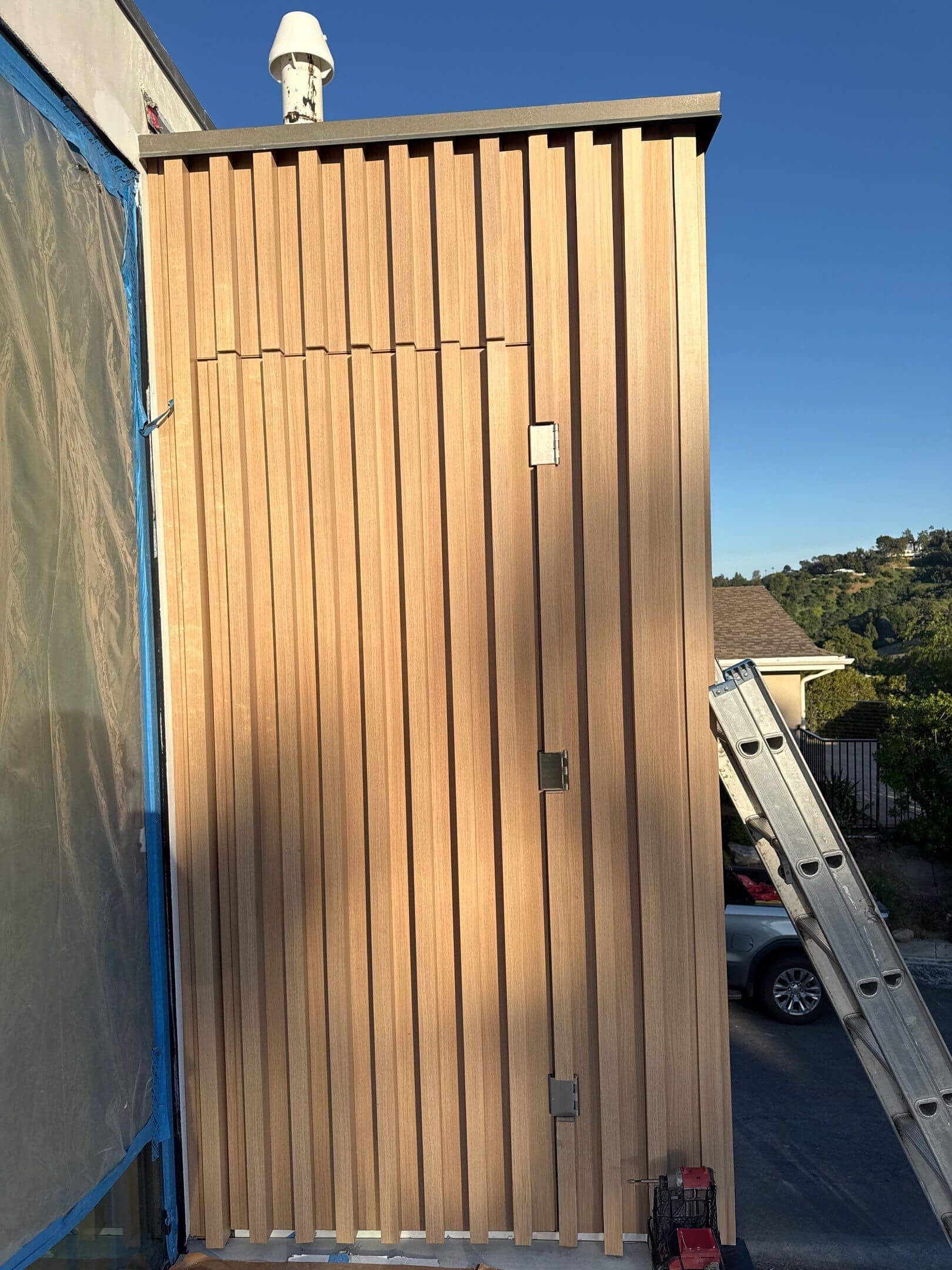 Vertical wood slats cladding the side of a building, with a silver ladder leaning against it and a pipe on the roof.