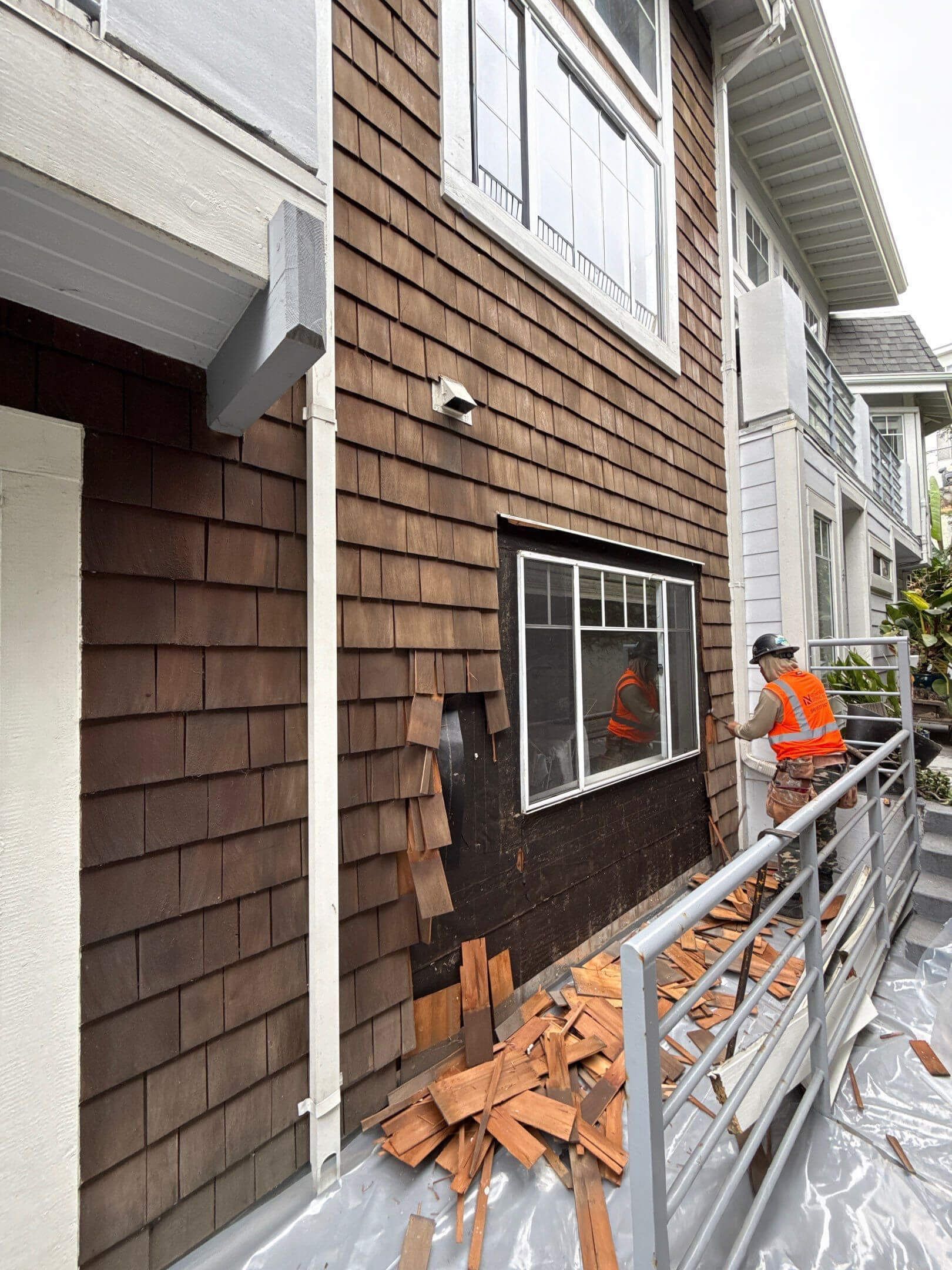 A construction worker in a high-visibility vest removes brown wooden siding from the exterior wall of a multi-story building.
