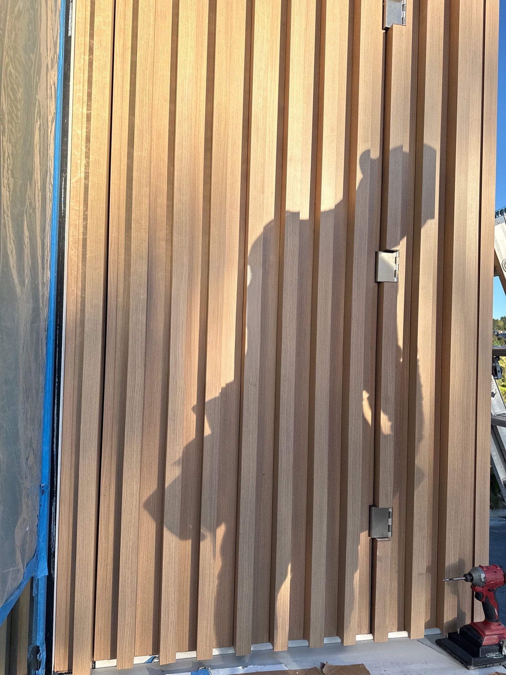Vertical wood slats are partially installed on a wall, with a power drill visible on the floor and a shadow cast on wood.