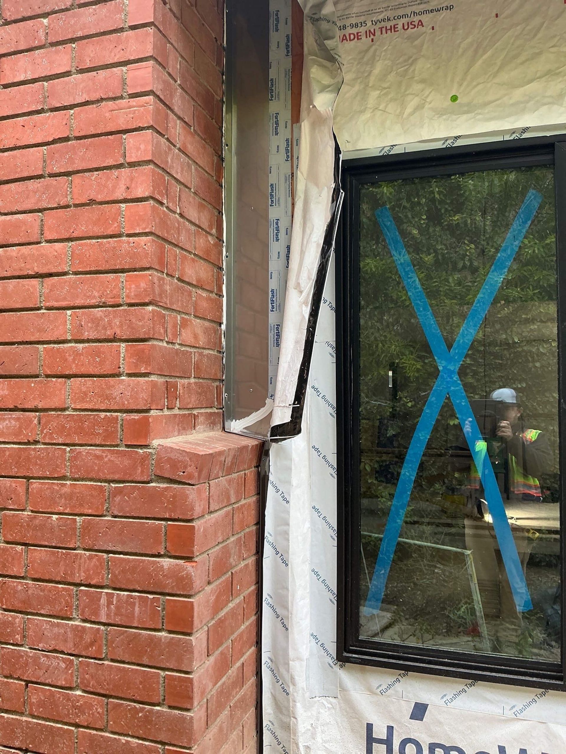 A partially installed window with blue tape on the glass sits next to a red brick wall with unfinished siding and trim.