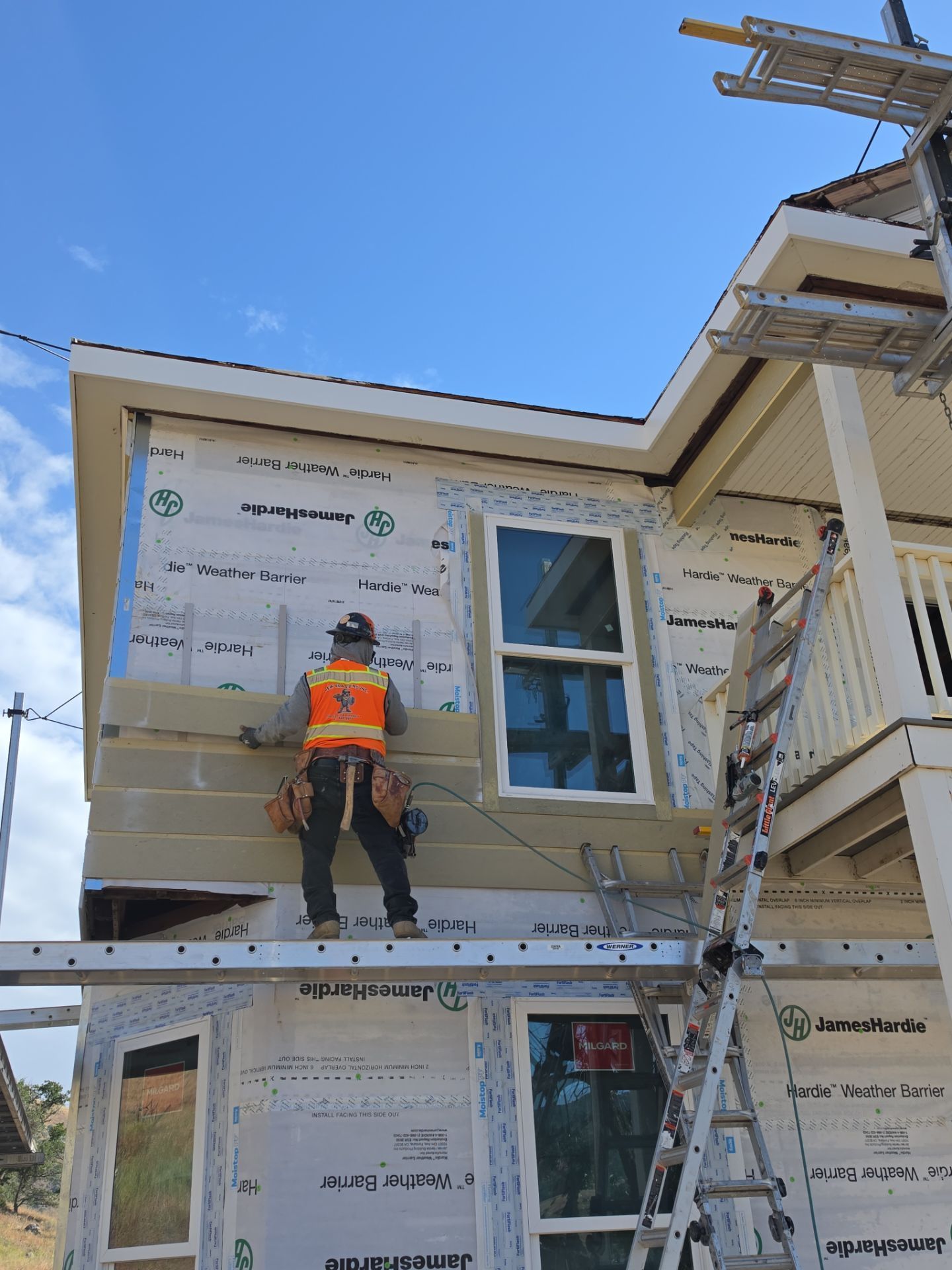 Worker in orange safety vest installing siding on a house exterior under a blue sky.