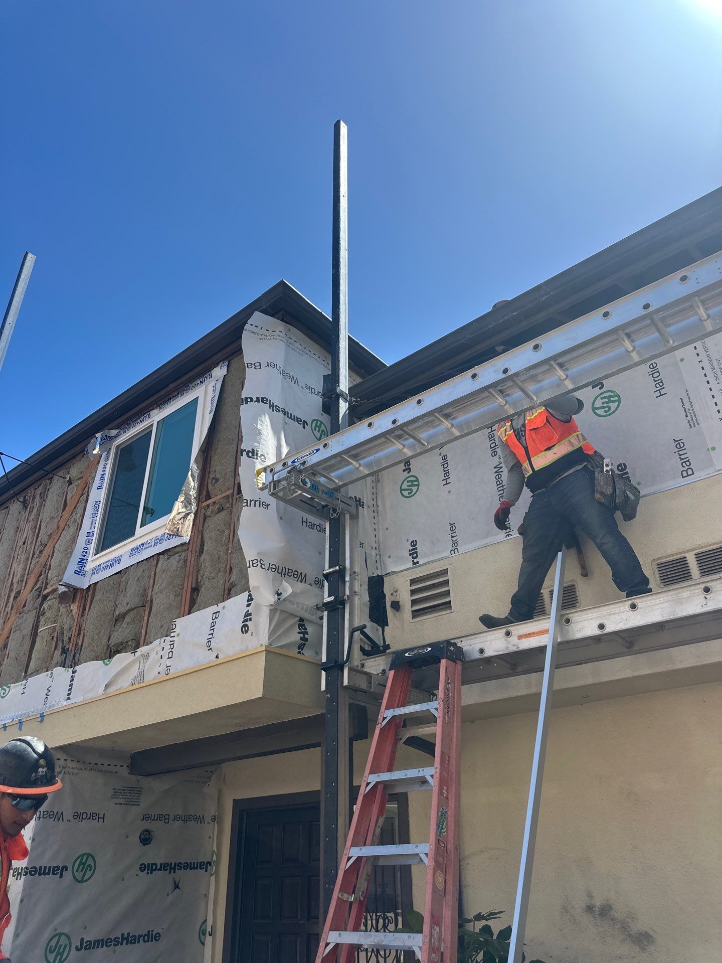 Construction workers on a house exterior, one on a ladder installing siding beneath a blue sky.