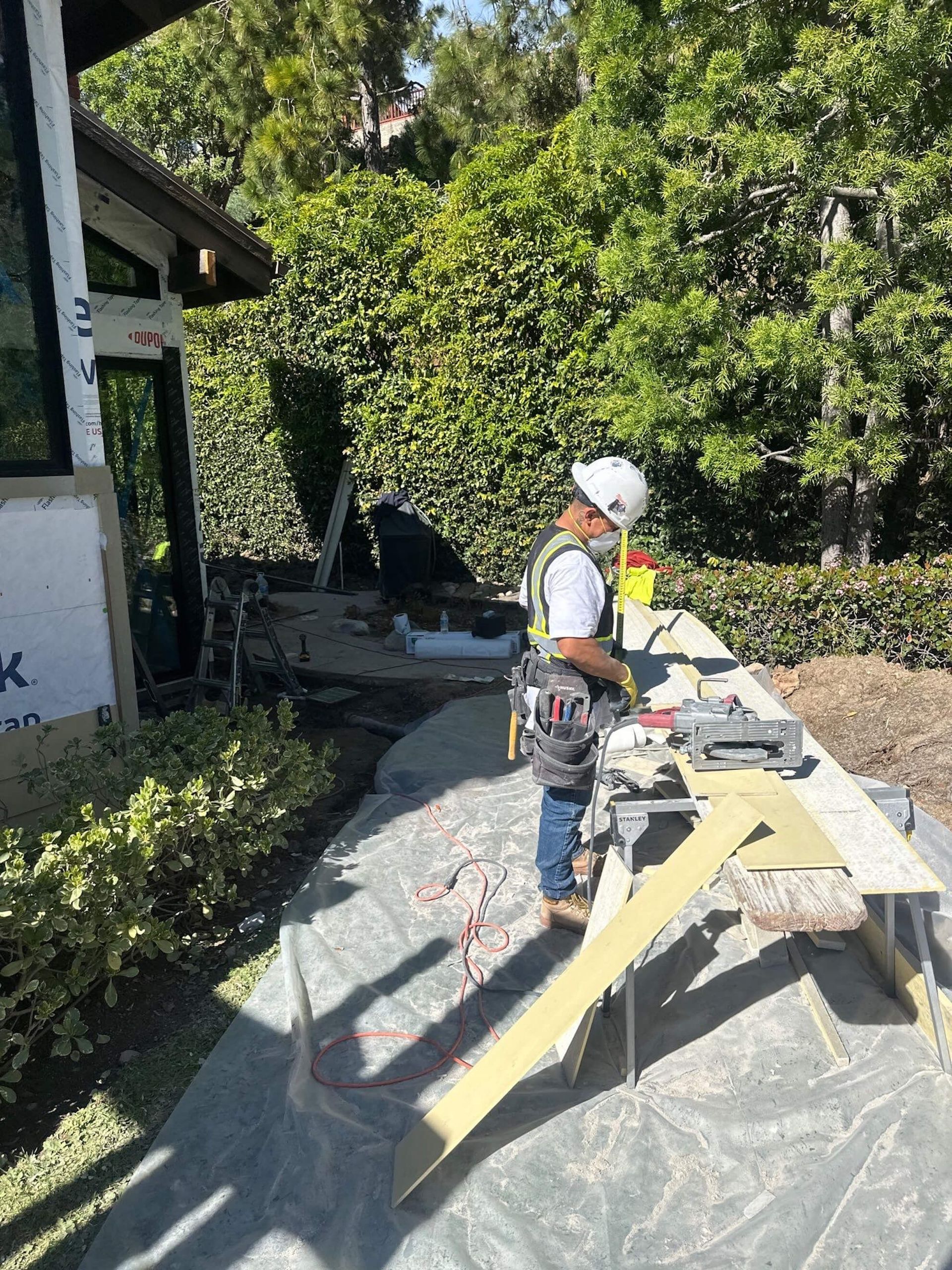 A construction worker in a white hard hat and safety vest uses a circular saw on a wooden plank at an outdoor job site.