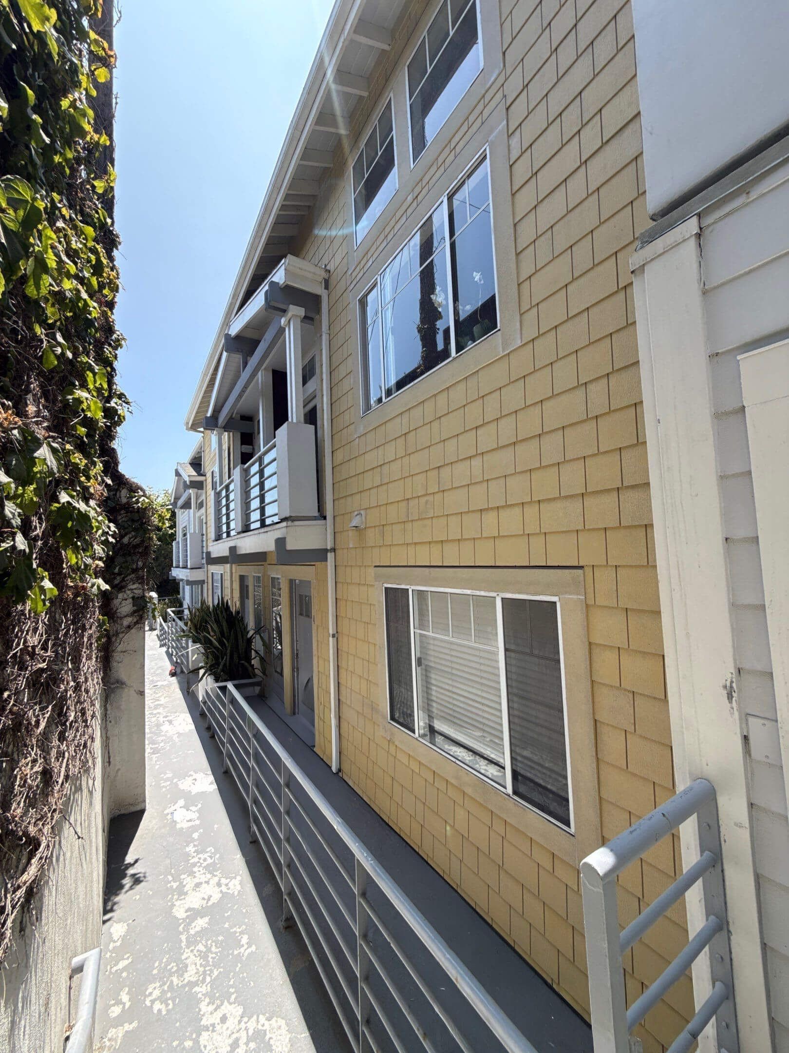 A narrow outdoor walkway runs along the side of a multi-story tan brick building with several windows and a metal railing.