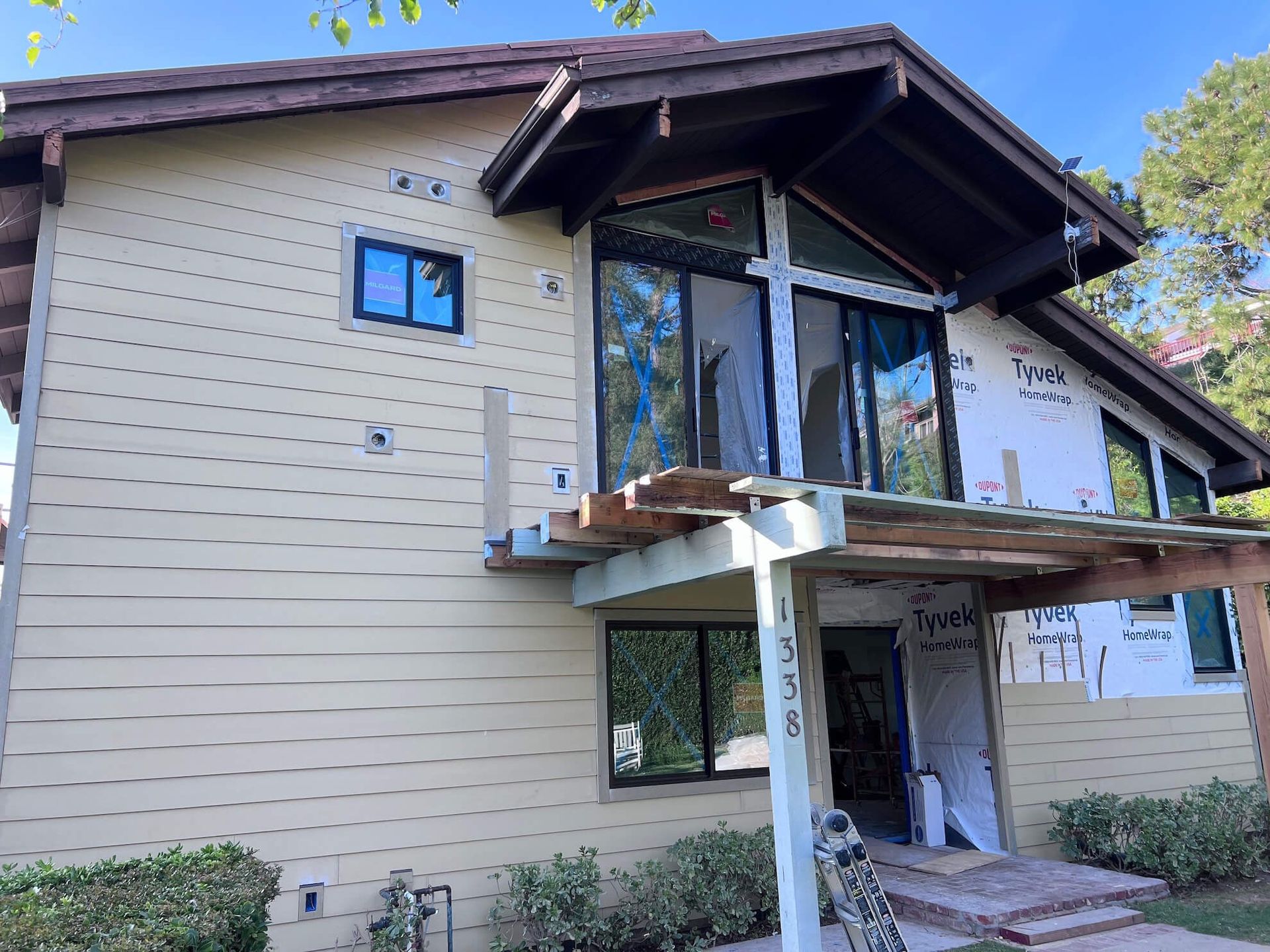 A tan two-story house under renovation, featuring exposed Tyvek house wrap, a balcony without railings, and large windows.