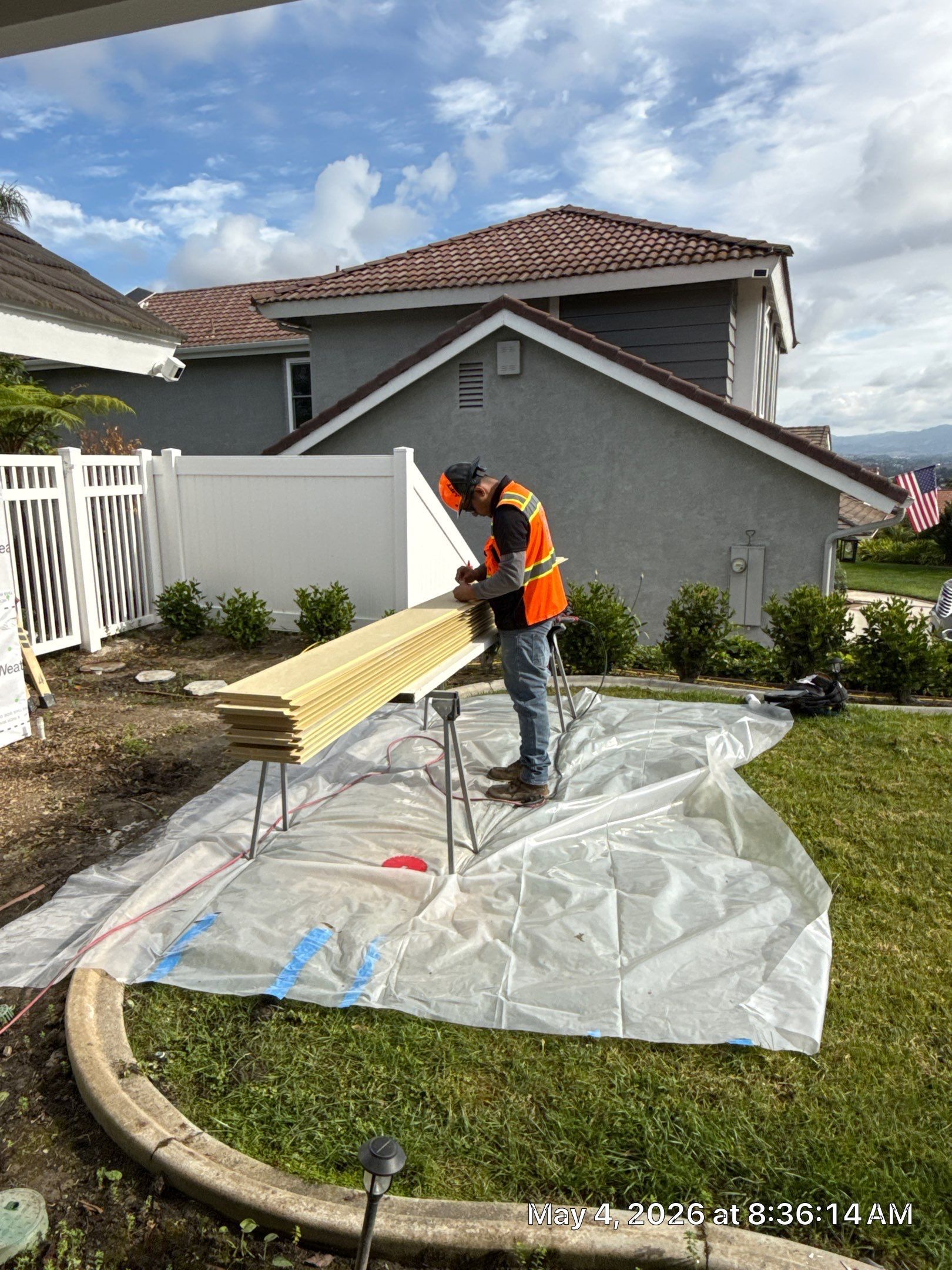 Worker in orange vest cutting wood on a tarp in a suburban yard beside a house