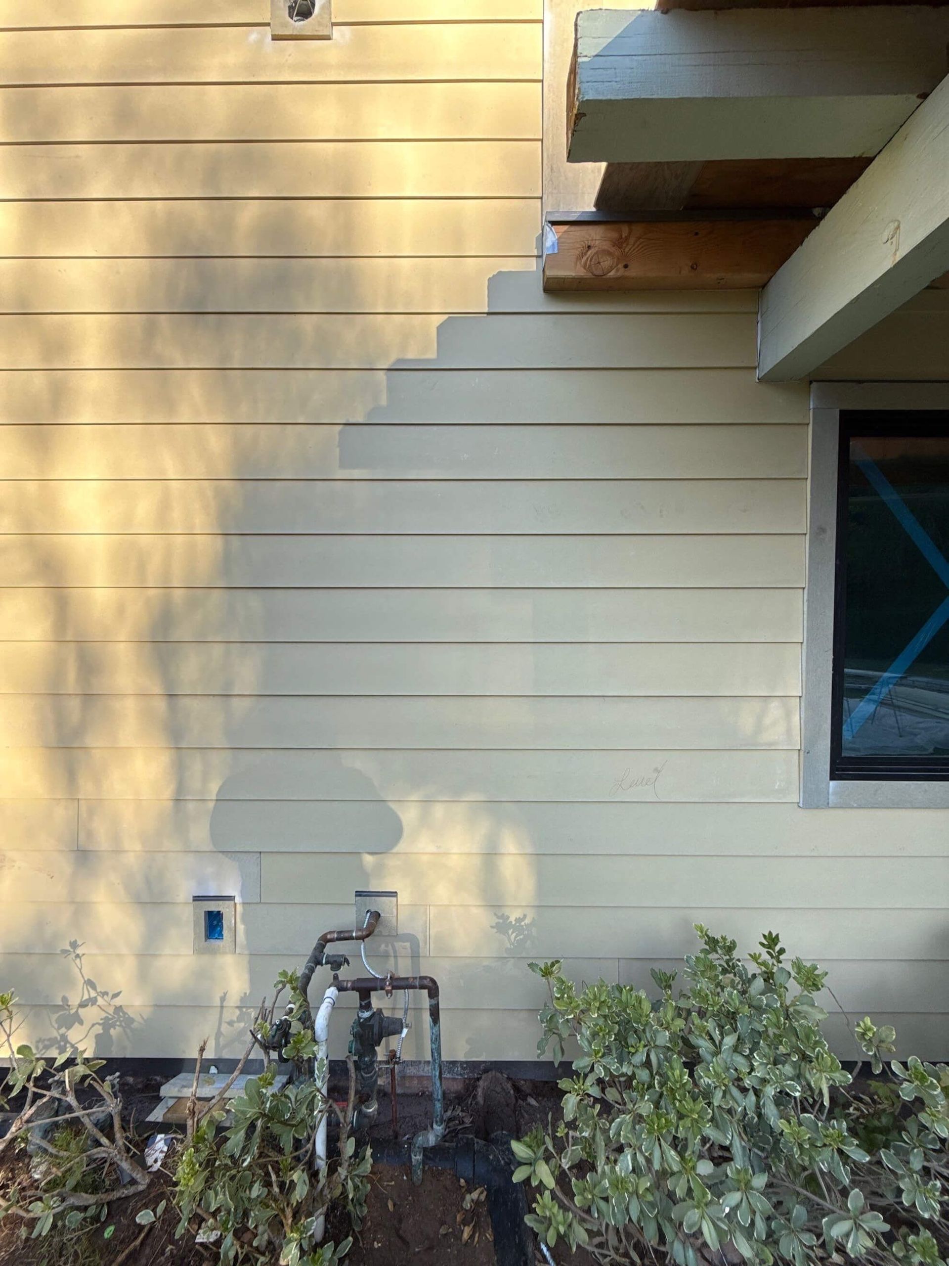 Exterior wall with horizontal beige siding, an exposed beam overhang, a window, and utility pipes near ground-level shrubs.