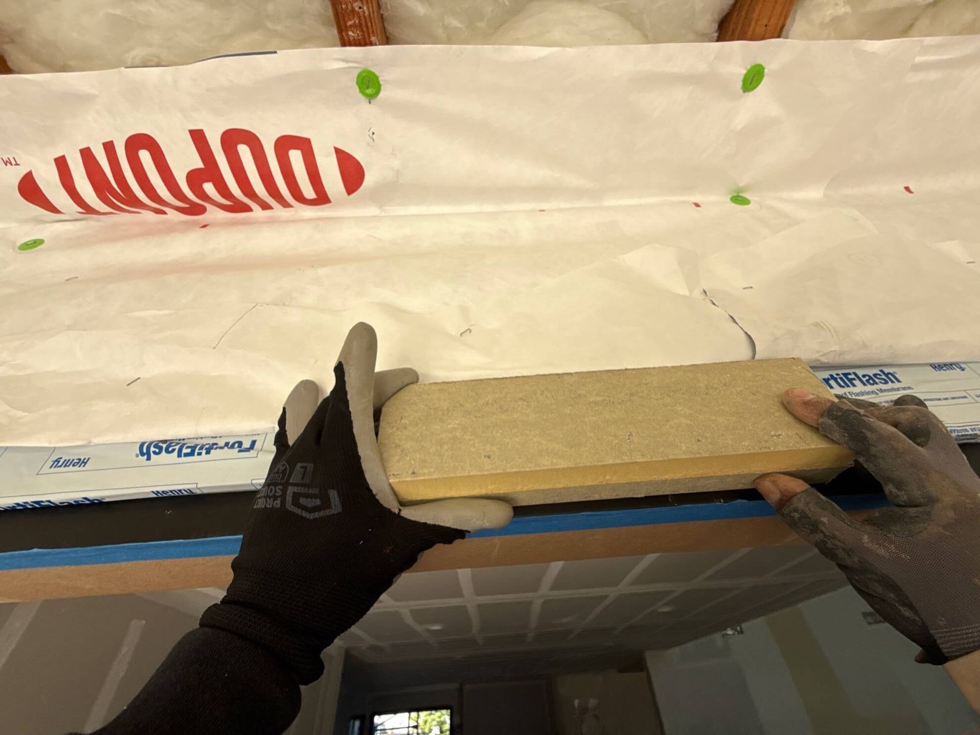 A first-person view of gloved hands holding a block of insulation against a white house wrap barrier in a wall frame.