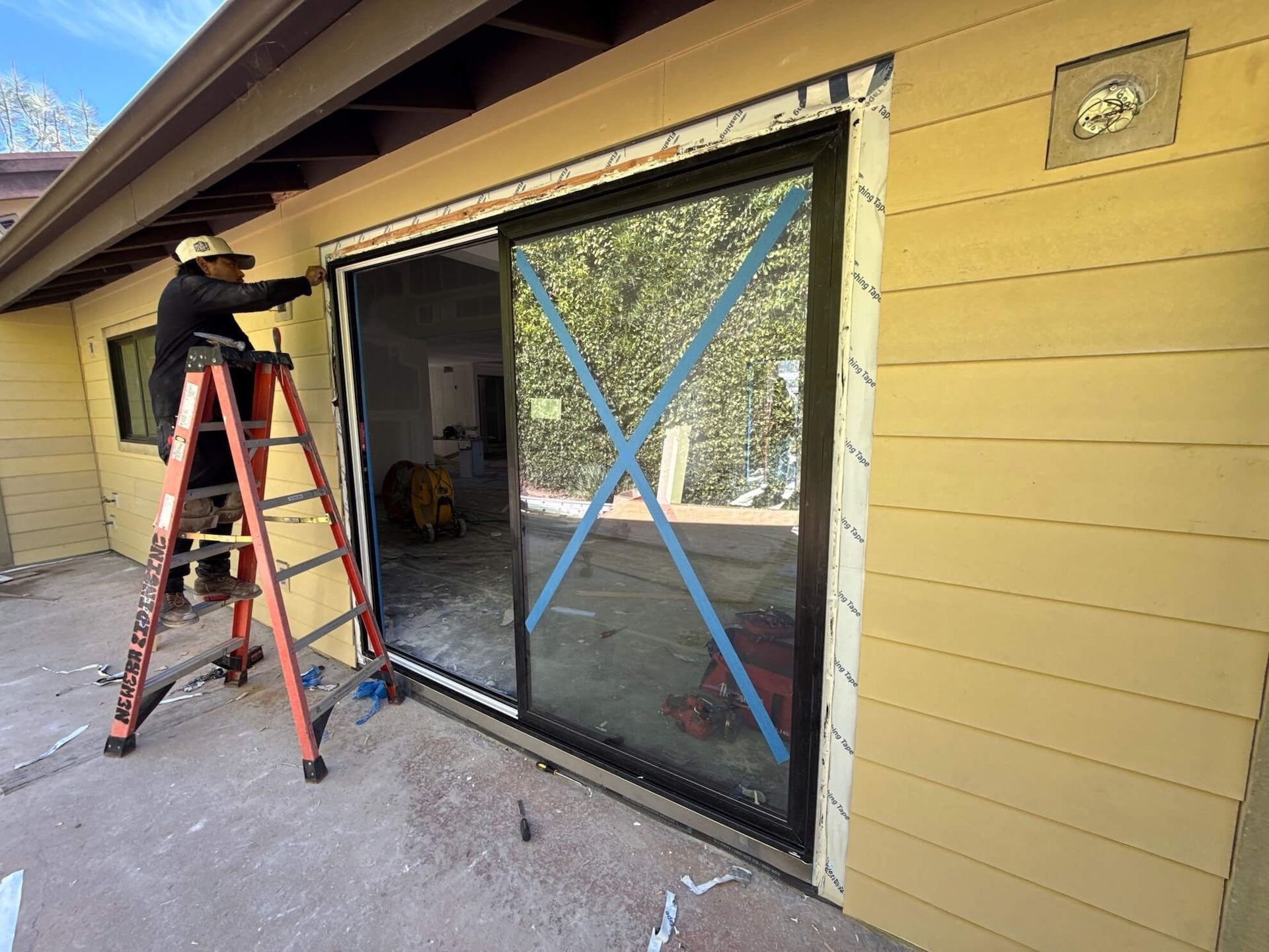 A worker on a ladder installs a sliding glass door on the side of a yellow-sided building.