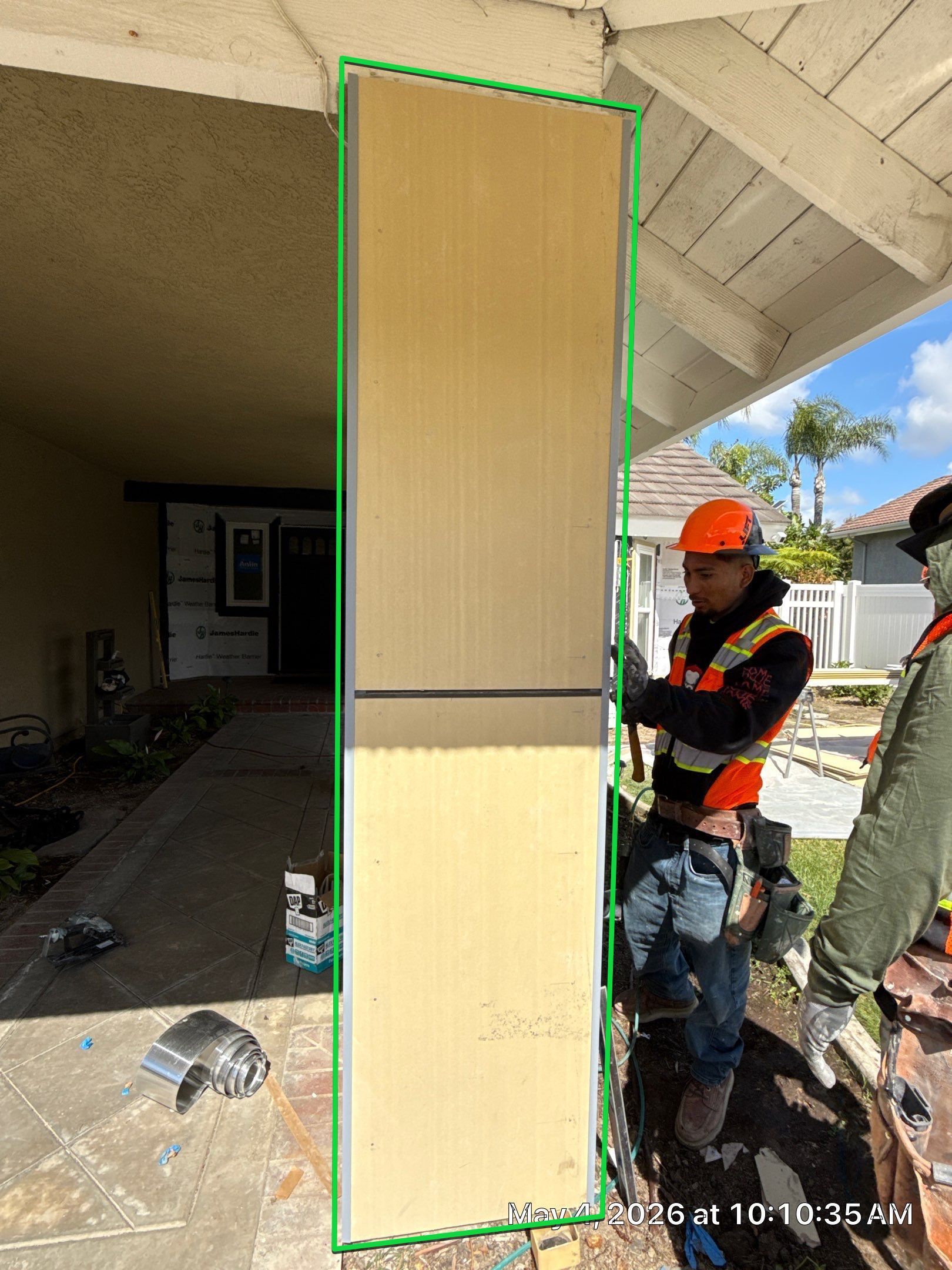 Worker in orange hard hat inspecting a tall beige panel under a covered patio.