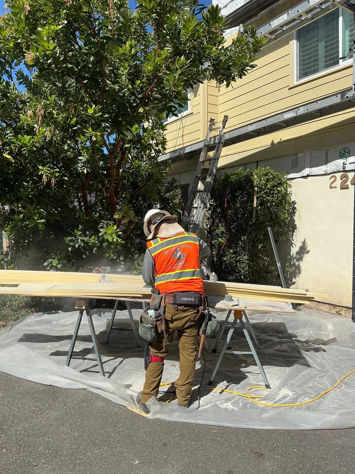Worker in orange safety vest cutting pavement on a driveway beside a beige building.