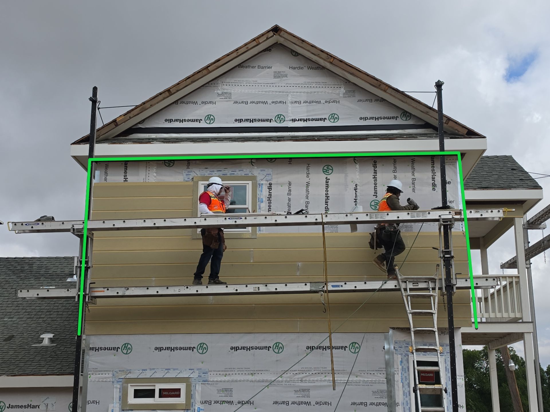 Workers on scaffolding installing siding on a house exterior under cloudy skies