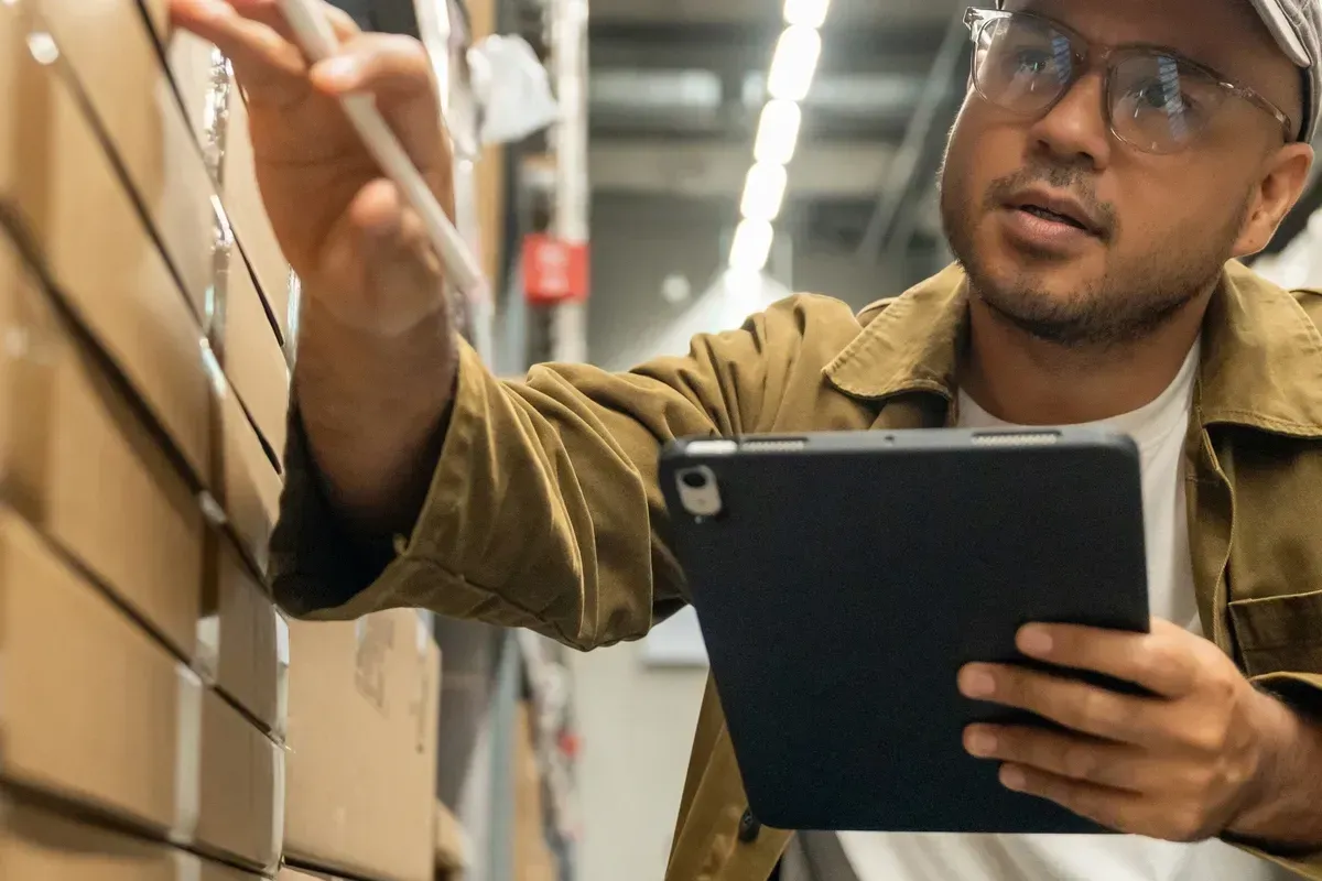 Worker in a warehouse checking inventory with a tablet beside stacked cardboard boxes
