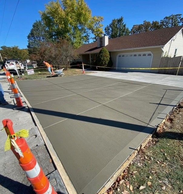 Freshly poured concrete driveway beside a house, with orange cones and caution tape blocking the work area.
