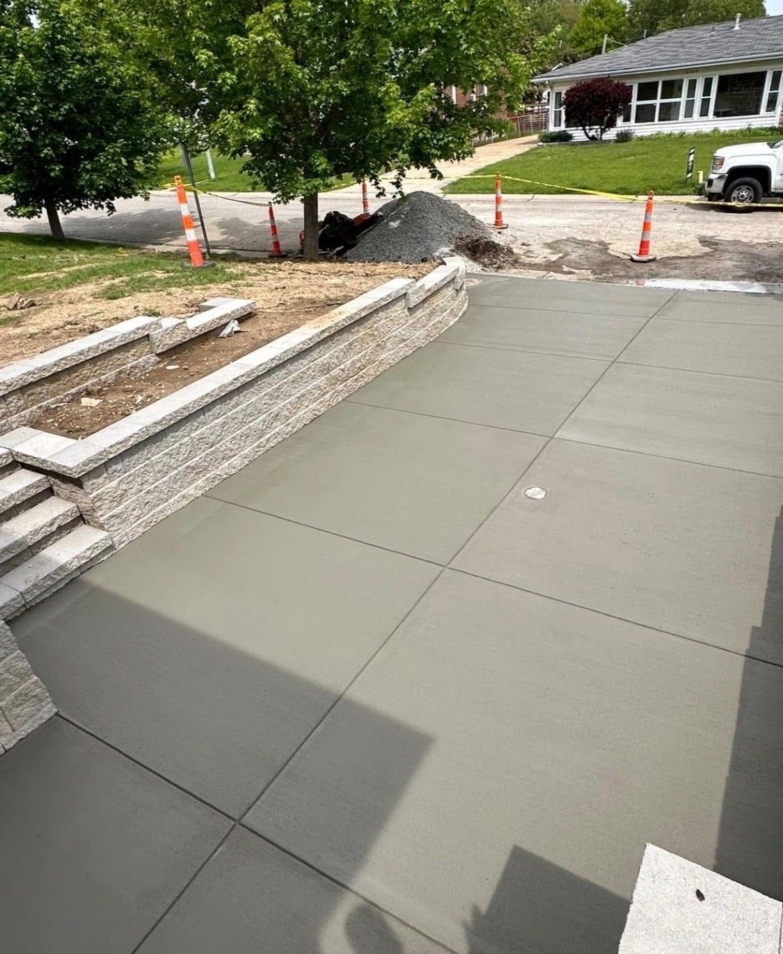 Fresh concrete patio beside a stone wall, with steps, cones, and a driveway in a residential yard