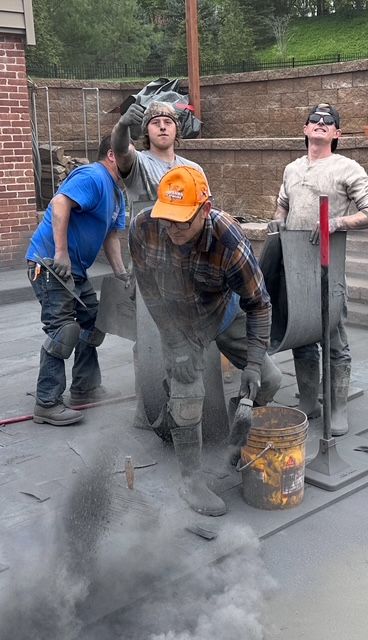 Three workers in hard hats and face shields lean over a smoking metal barrel outdoors