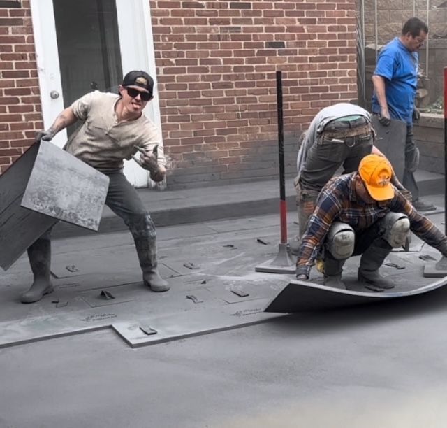 Workers installing a roof membrane, one person kneeling over gray sheets, another carrying material by a brick wall.