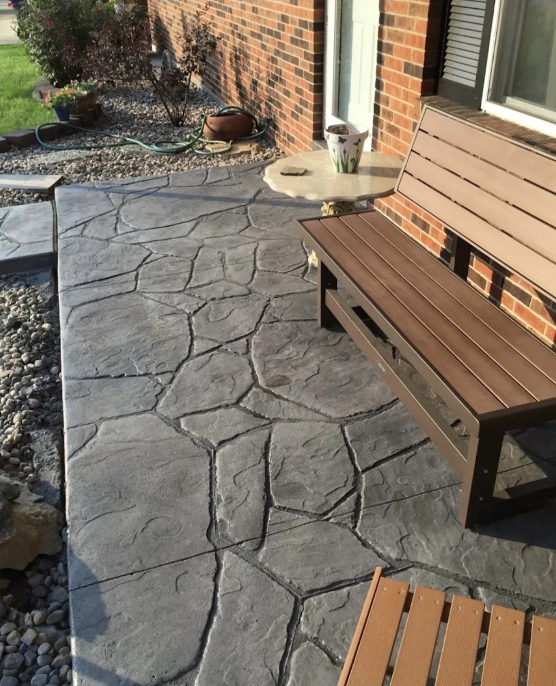 Stamped concrete patio beside a brick house, with wooden bench and planters by the door.