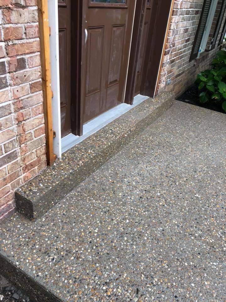 Open front door with a low step down to a pebble-textured patio beside a brick wall