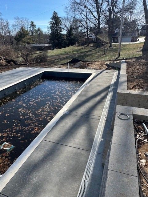 Concrete spillway beside a small pond, with leaf debris and trees in a park-like setting.