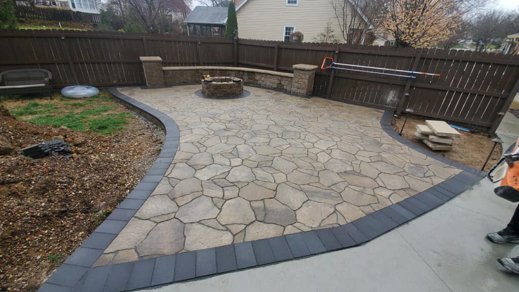 Backyard patio with stone pavers, curved border, and a wooden fence; person partly visible at right.