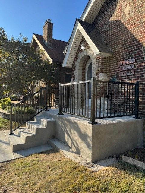 Brick house entrance with concrete steps, black railing, and arched doorway in sunlight