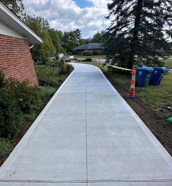 Curved concrete sidewalk beside a brick building, with landscaping and trash bins nearby.