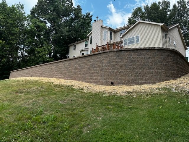 Large house with a brick retaining wall on a grassy hillside under a partly cloudy sky
