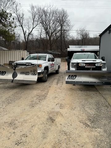 Two white snowplow trucks parked on a gravel lot in front of bare trees.