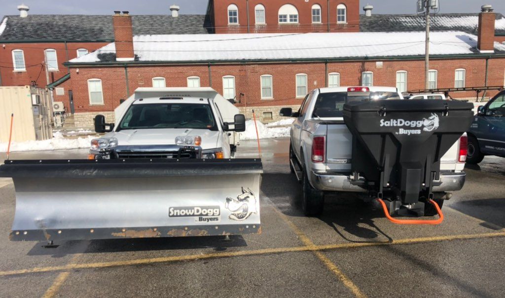 Two trucks parked in a lot, one gray with a snowplow and one black with a salt spreader