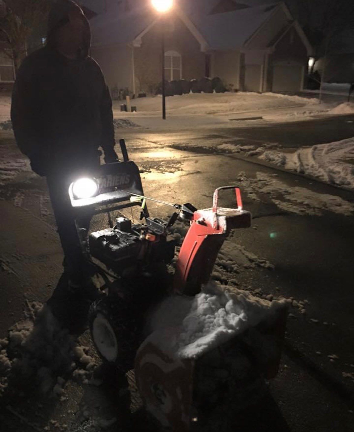 Person standing by a snowblower clearing snow at night on a snowy street under a streetlight