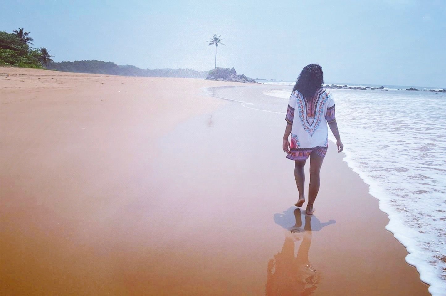 Jess walks barefoot along a sandy beach near the ocean while wearing a white patterned cover up.