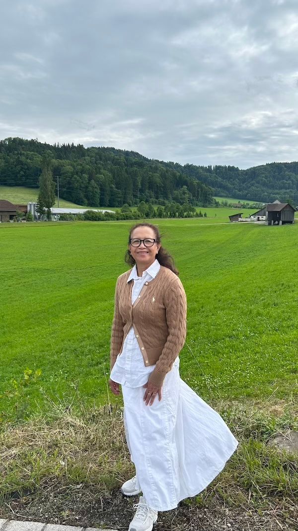 A smiling grandmother stands in a bright green field in the Appenzell countryside with forested hills. The peaceful rural scenery shows why Switzerland is worth visiting for quiet landscapes and village views.