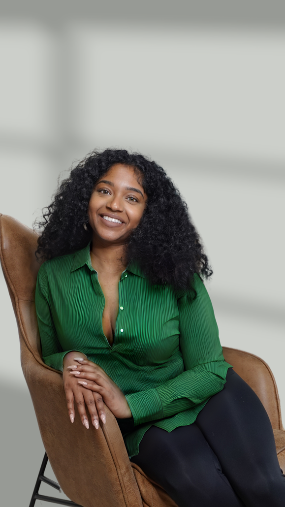 Jess sits in a brown leather chair against a soft gray background. She has curly black hair and is smiling while wearing a green blouse.