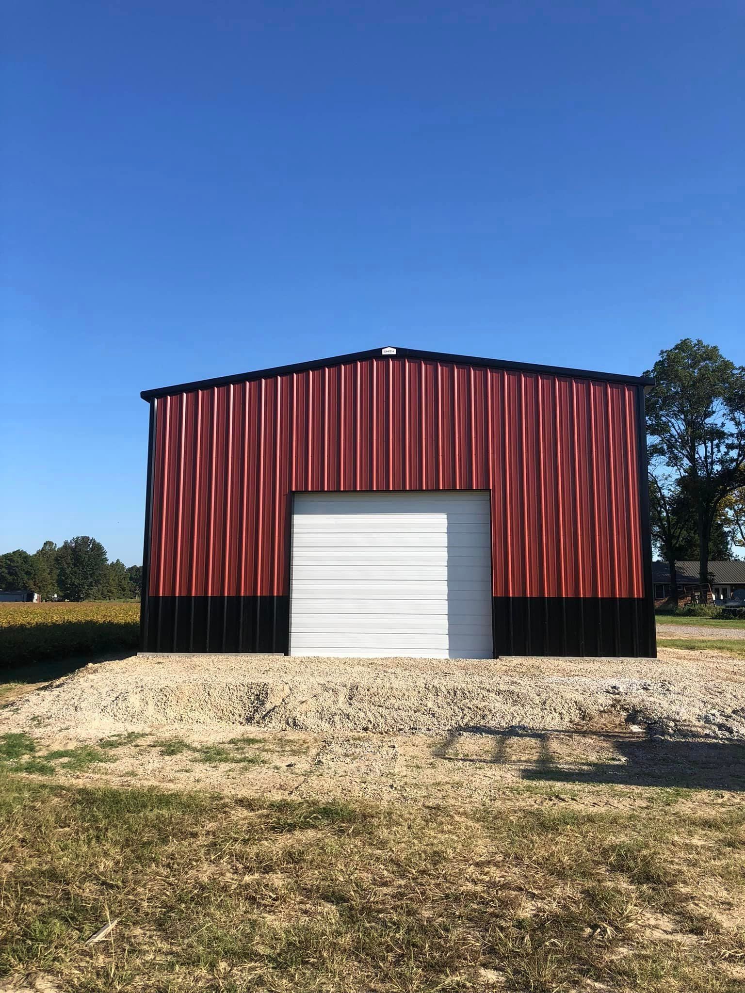 A red and black barn with a white door is sitting in the middle of a field.