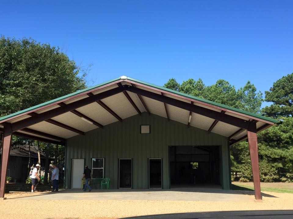 A green open-sided metal pole barn with a gabled roof, standing on a gravel lot under a clear blue sky.