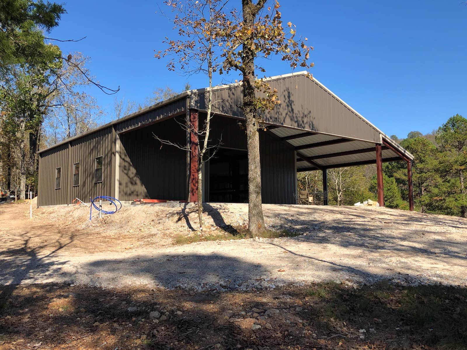 A brown metal pole barn with a covered porch sits on a gravel lot surrounded by trees under a clear blue sky.