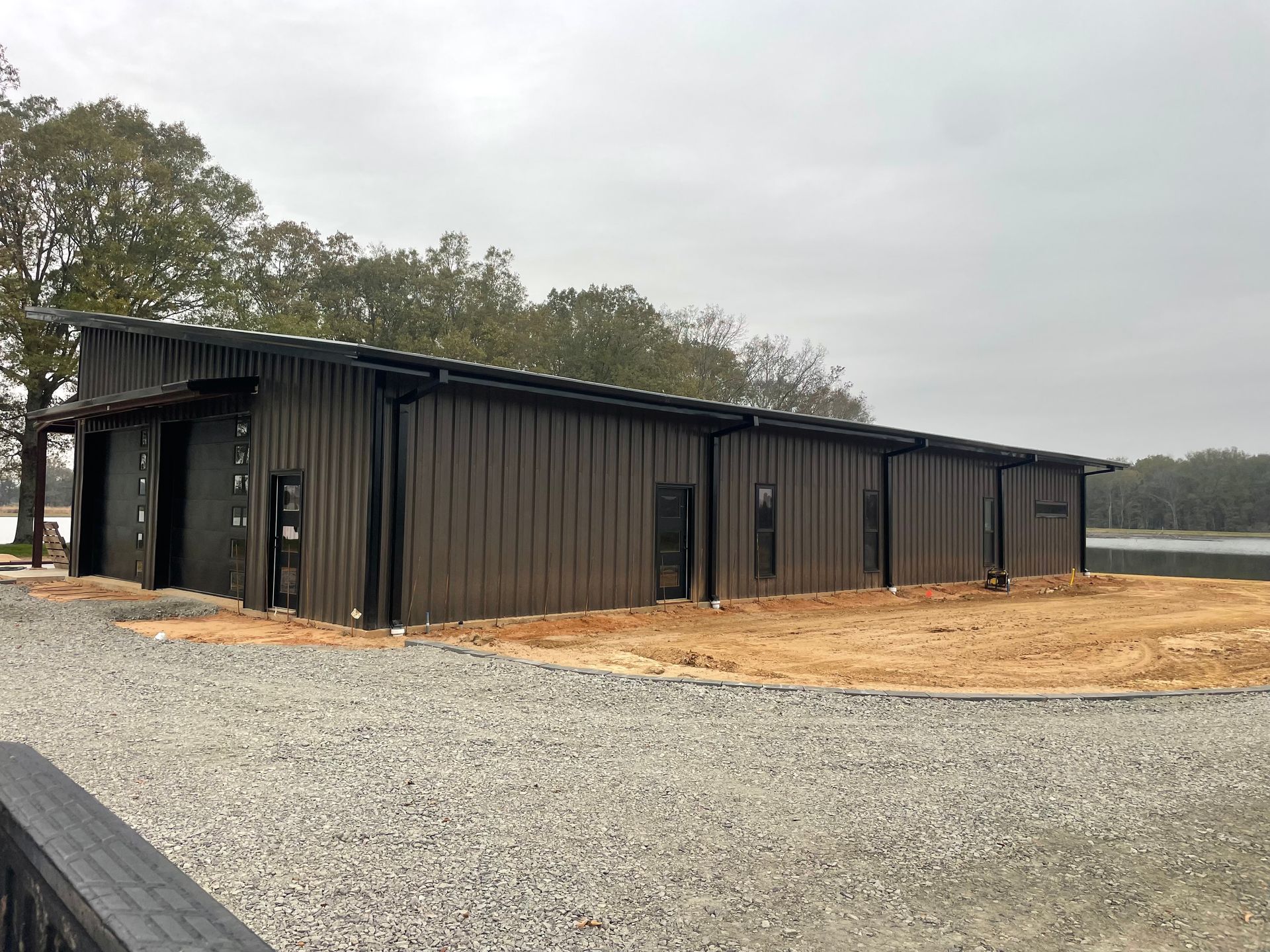 A modern, dark-brown metal building with a sloped roof sits on a gravel lot near a line of trees and a body of water.