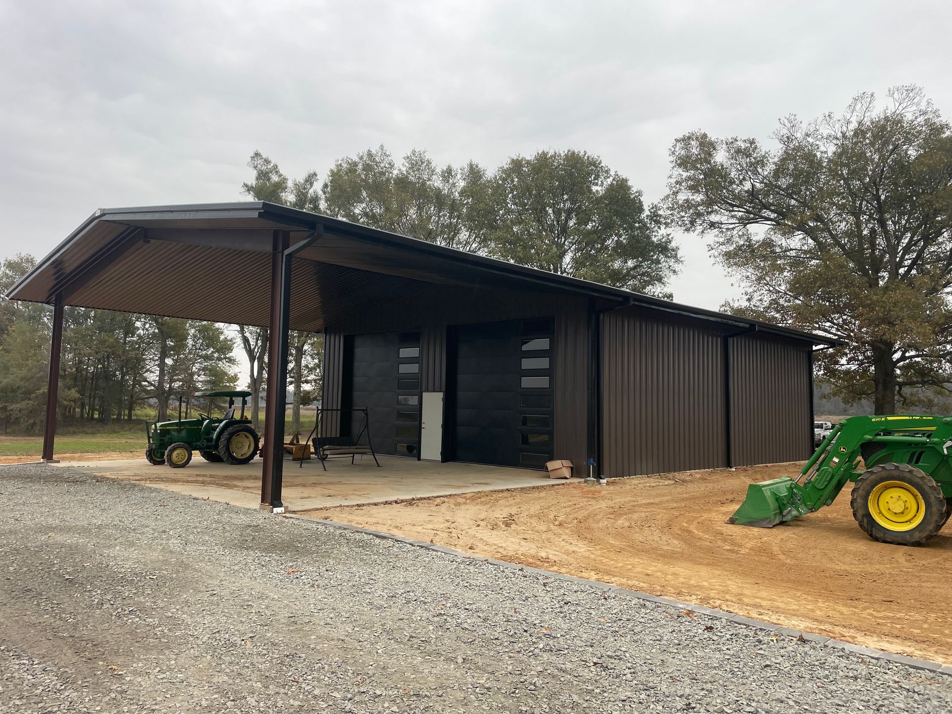 A dark metal barn with an open carport area and two garage doors, featuring green tractors parked on a gravel lot.