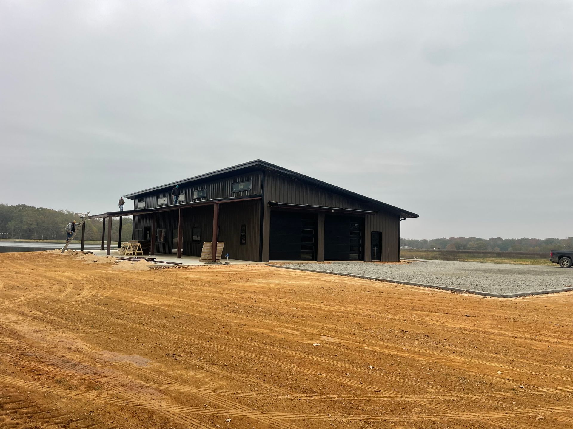 A dark-colored, modern barn-style building with a metal roof, situated on a large, cleared lot of dirt and gravel.
