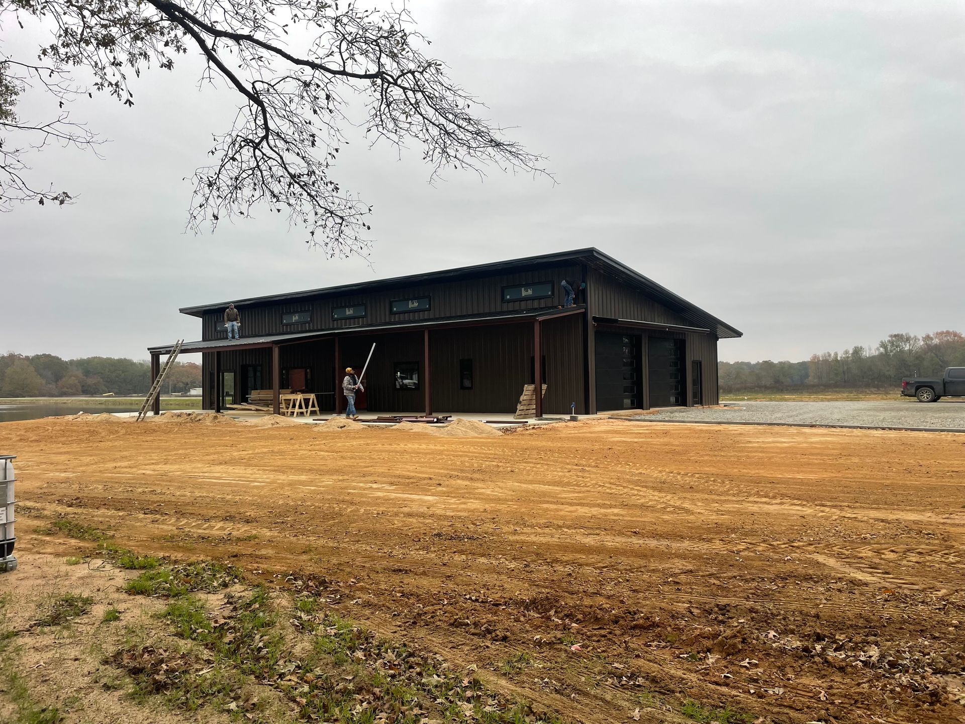 A dark-sided building with a covered porch and garage bays sits on a construction site with exposed dirt under a gray sky.