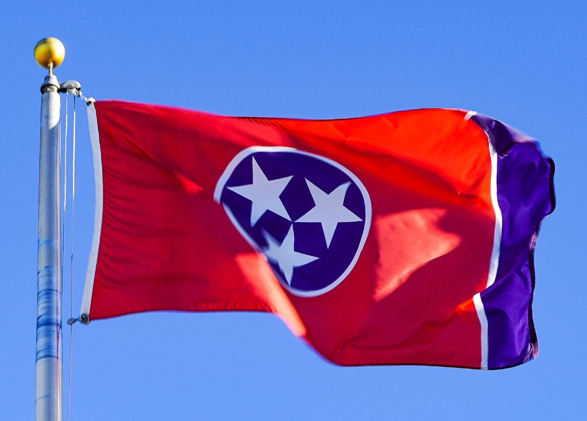 The Tennessee state flag, a red field with three white stars in a blue circle and a vertical blue stripe, waving against sky.