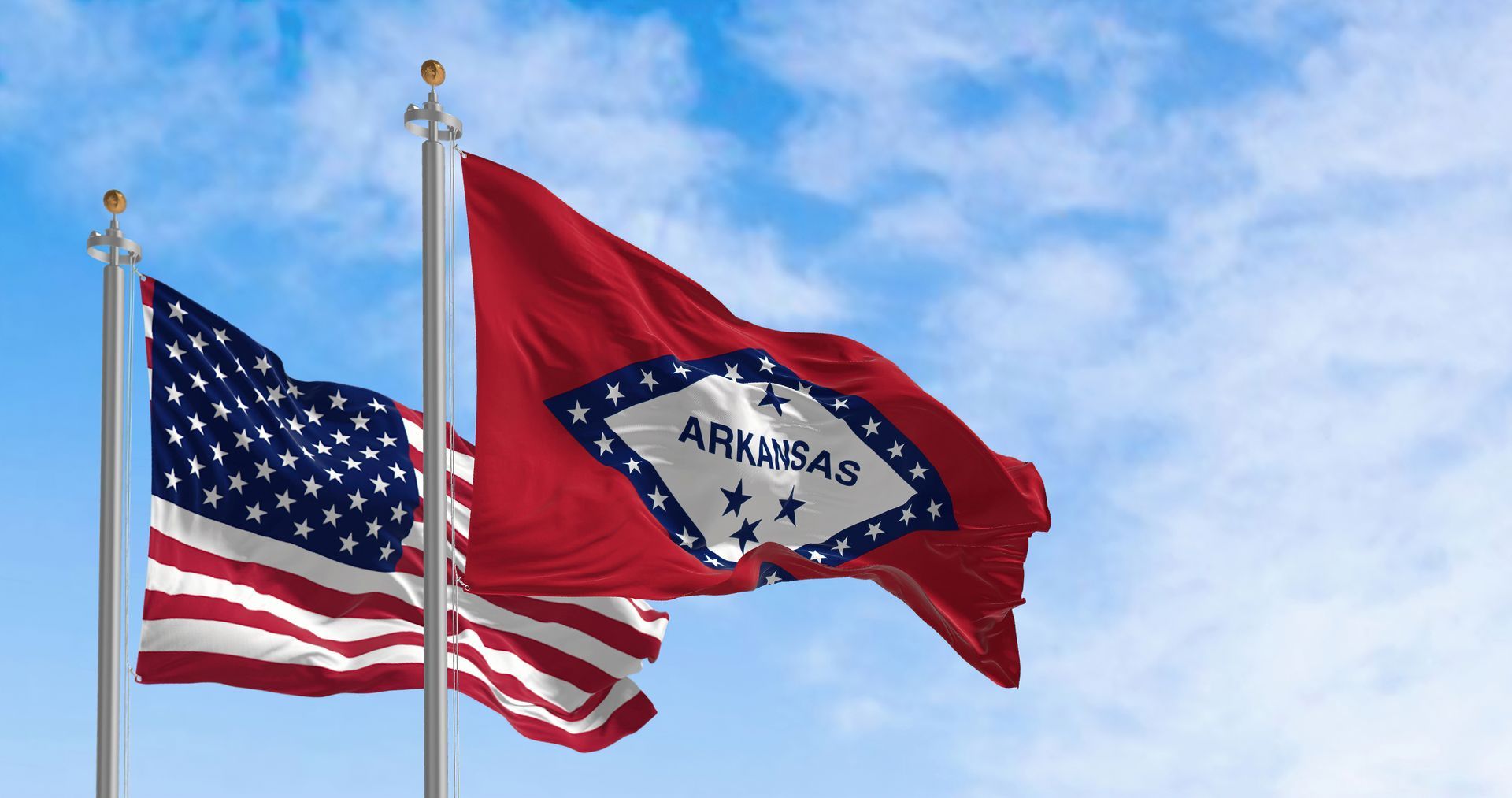 The US flag and the Arkansas state flag fly side-by-side against a blue, cloudy sky.