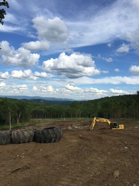 A yellow excavator is in a dirt field with mountains in the background