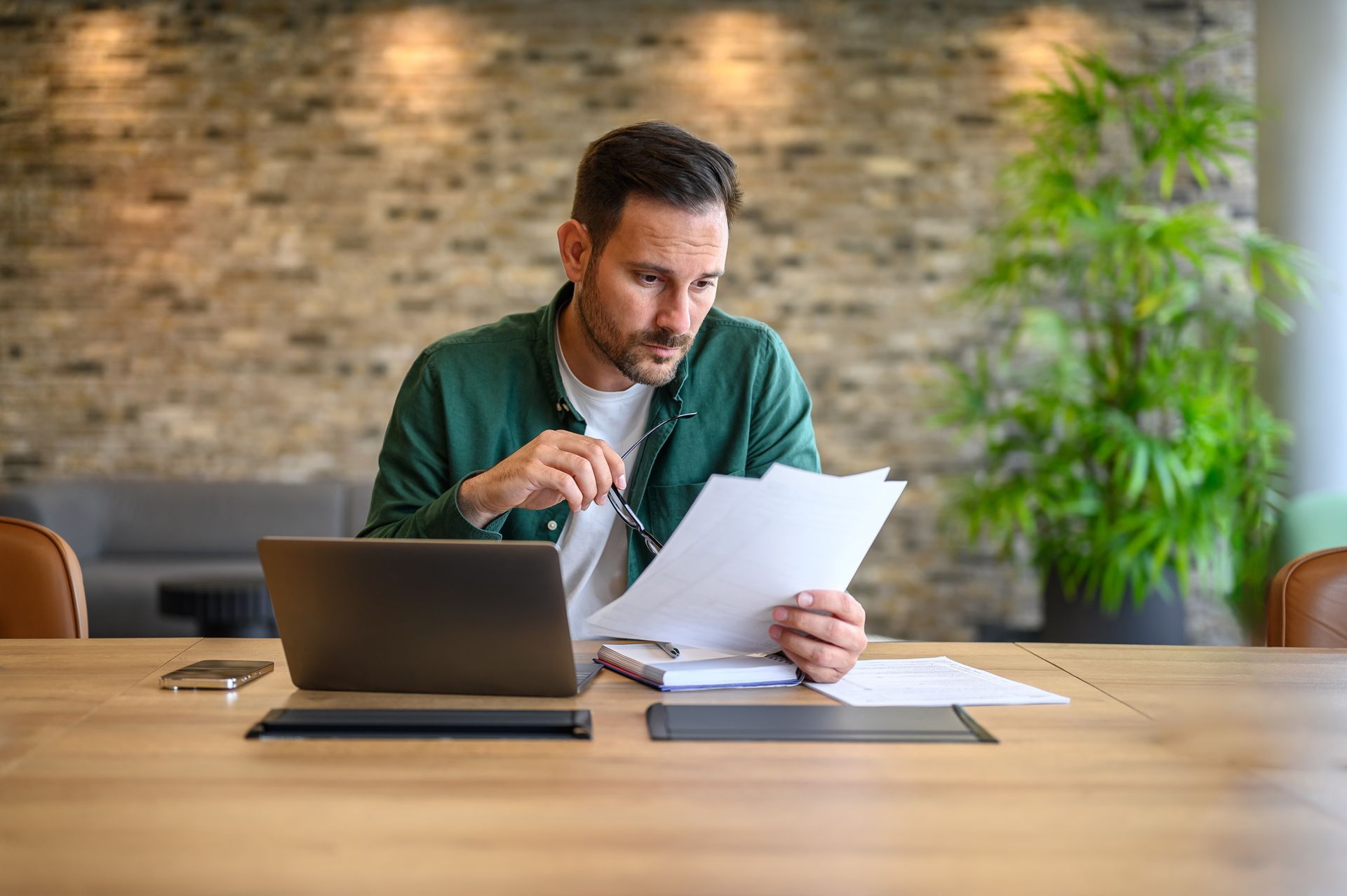 An accountant holding glasses and analysing a financial report with a laptop on a desk in an office.