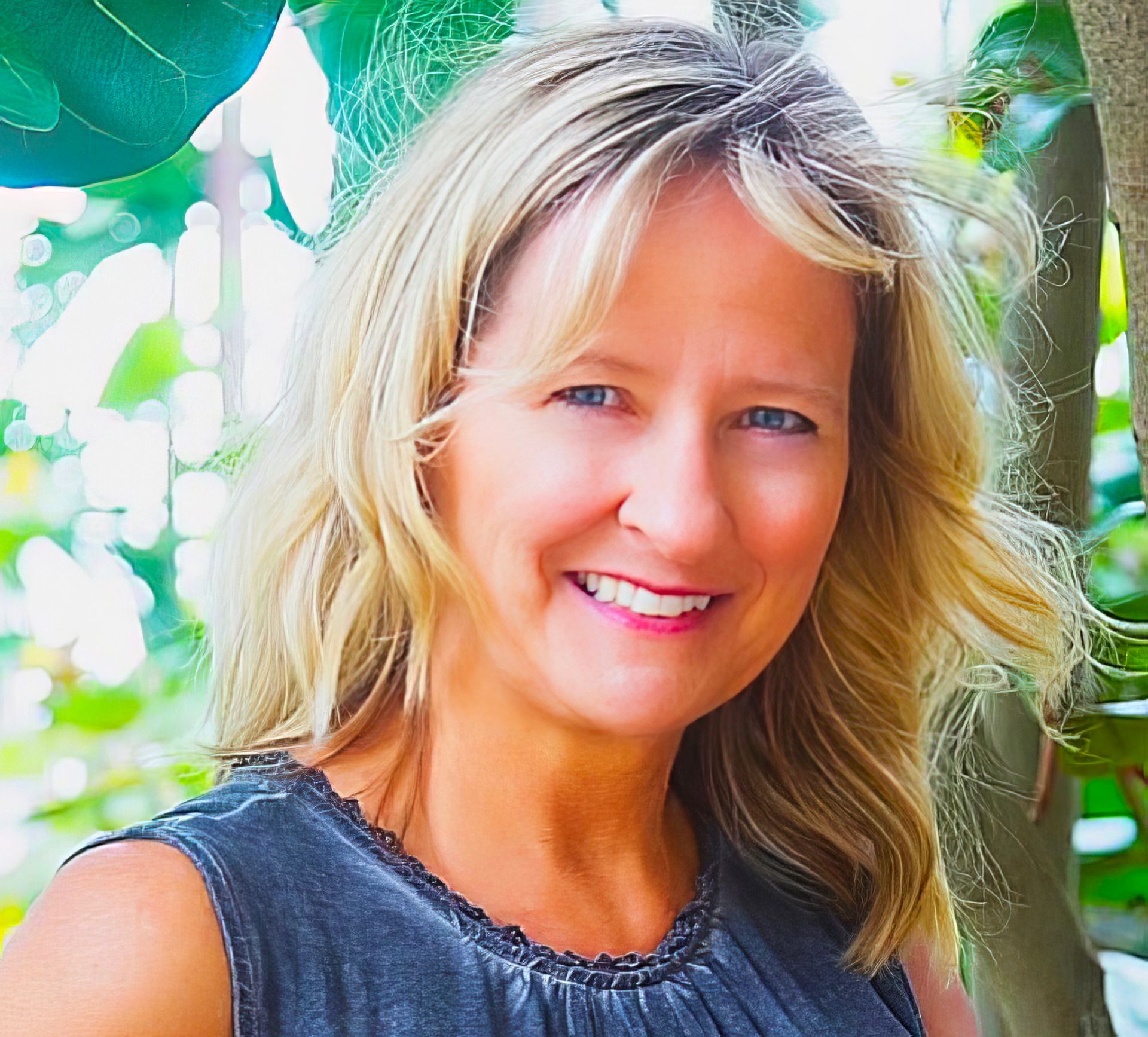 Blonde woman smiles, facing forward, in outdoor setting, wearing gray sleeveless shirt.
