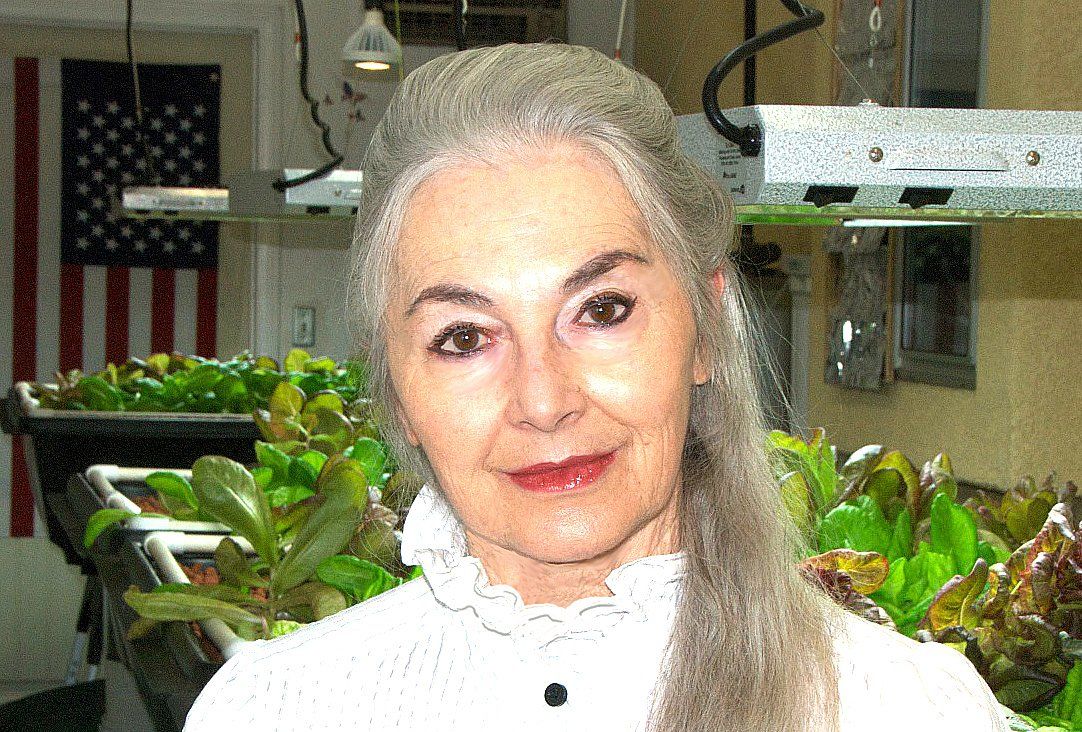 Woman with grey hair stands by hydroponic lettuce, American flag in background.
