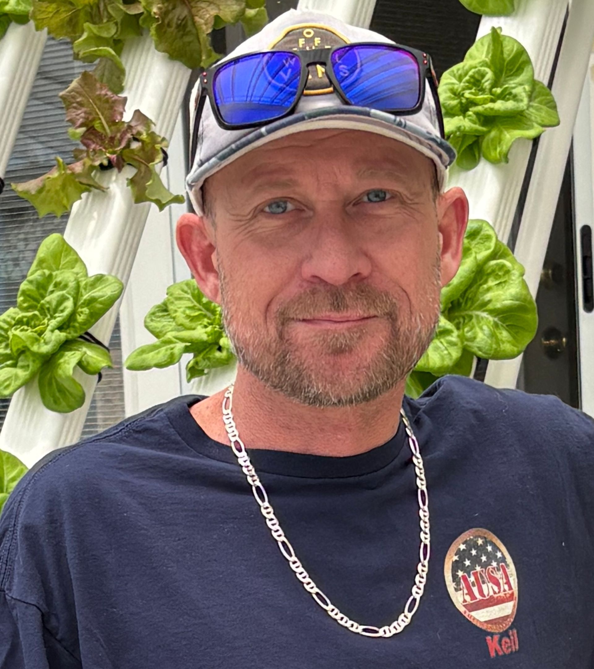 Man in blue shirt and hat, smiling, with lettuce plants in the background.